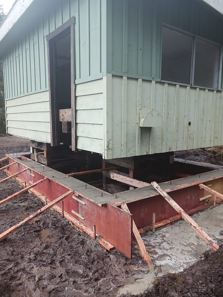 The small building on temporary supports within the new foundation forms, door open