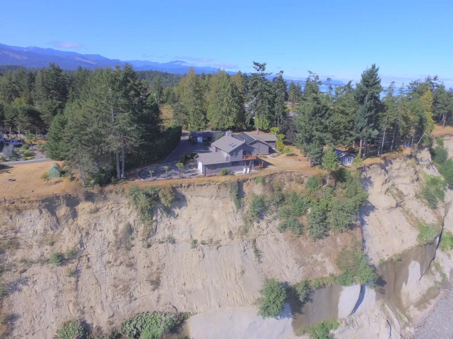 Aerial view of a high-bank waterfront property on the Olympic Peninsula, the house set close to the edge of a retreating bluff above the water.