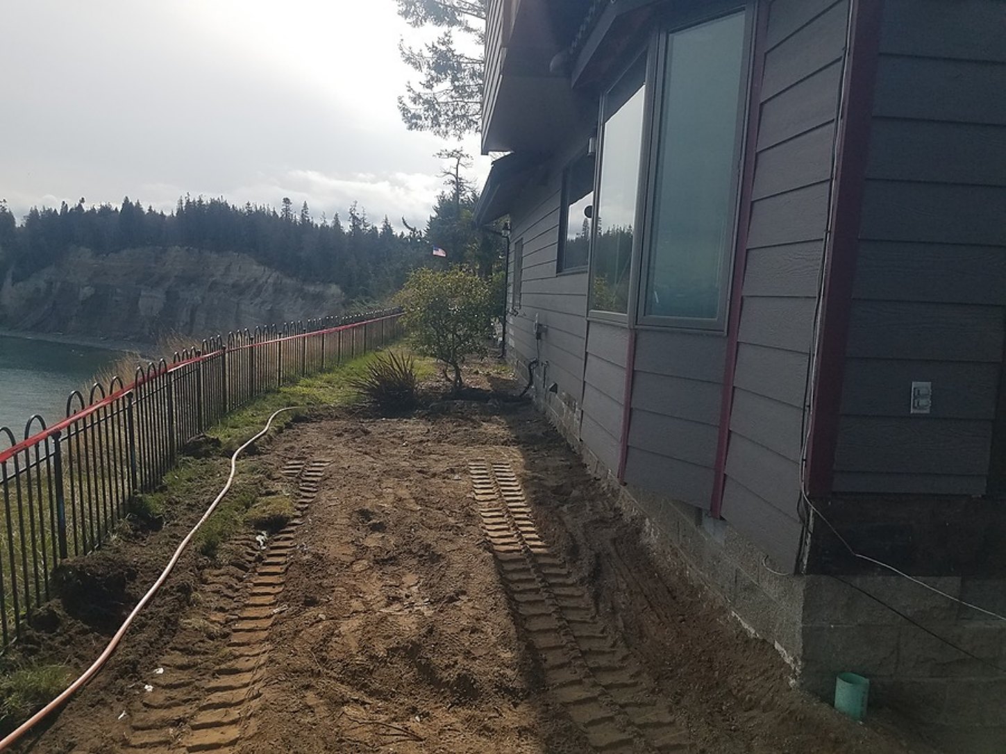A narrow strip of ground between a waterfront house and the edge of an eroding bluff, marked with excavator tracks, photographed before the house was relocated.