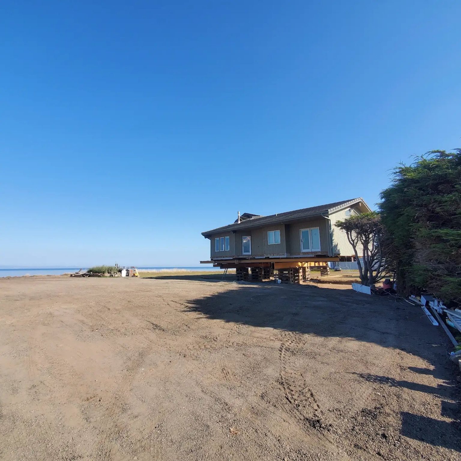 A relocated house standing on timber cribbing on open ground near the water, waiting for its new foundation.