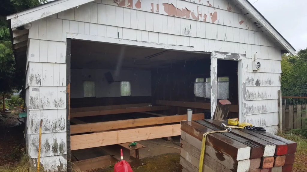A 1920s Port Angeles garage with peeling clapboard siding before repair, steel beams already set beneath the structure